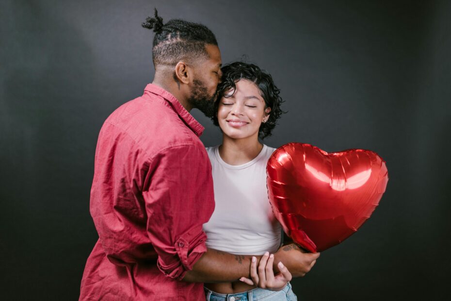 Ein glückliches Paar in inniger Umarmung, wobei der Mann einen Kuss auf die Stirn der Frau gibt, die ihre Augen geschlossen hat und ein strahlendes Lächeln zeigt. Sie halten gemeinsam einen roten herzförmigen Luftballon, was ein Gefühl von Romantik und Liebe in einem warmen und herzlichen Moment einfängt