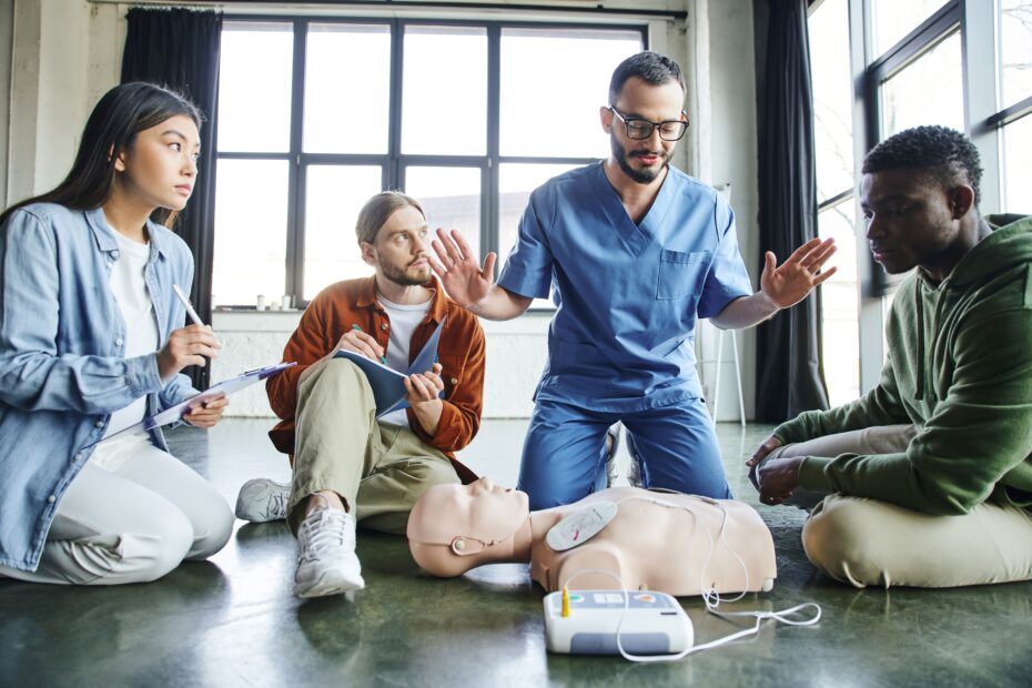 Eine Gruppe von Personen, die während einer Erste-Hilfe-Schulung von einem Fachmann lernen, wie man einen AED Defibrillator an einer Übungspuppe verwendet