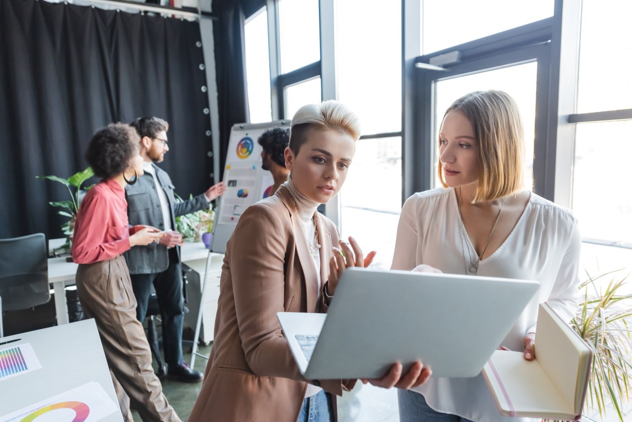 Zwei Marketing-Spezialistinnen diskutieren an einem Laptop, während im Hintergrund ein Team Strategien für eine Ghostwriting Agentur und Marketing-Kampagnen plant.