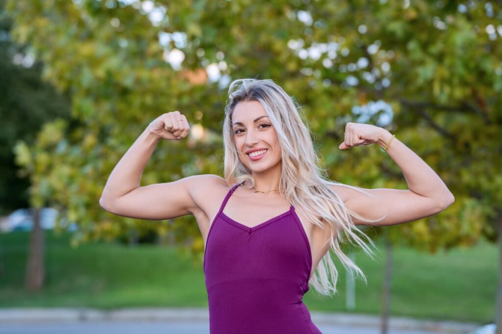 Frau zeigt stolz ihre trainierten Arme in einem Park – Symbol für einen erfolgreichen Start ins neue Jahr mit Unterstützung eines Personal Trainers.